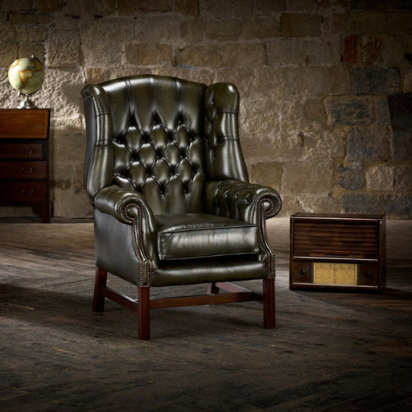 A green upholstered Georgian wing chair with padded wings, sides, and arms, featuring a polished wooden 'H' shaped sub-frame, situated in a room with a stone wall.
