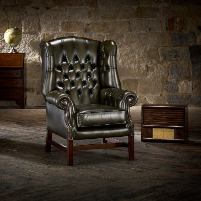 A green upholstered Georgian wing chair with padded wings, sides, and arms, featuring a polished wooden 'H' shaped sub-frame, situated in a room with a stone wall.